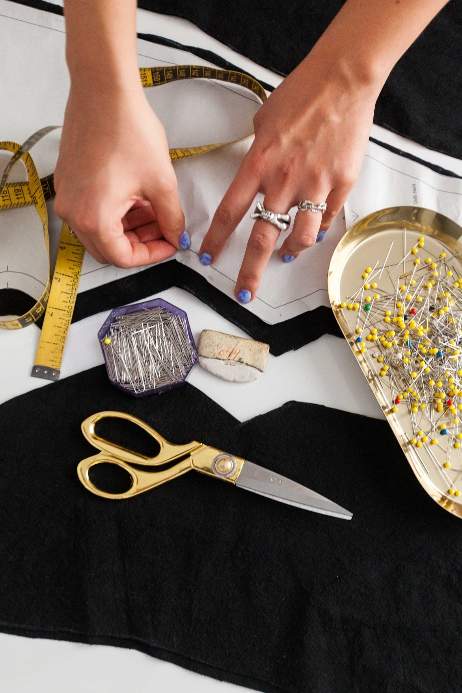 Hands pinning a paper pattern to fabric beside a tape measure and gold scissors on a white table