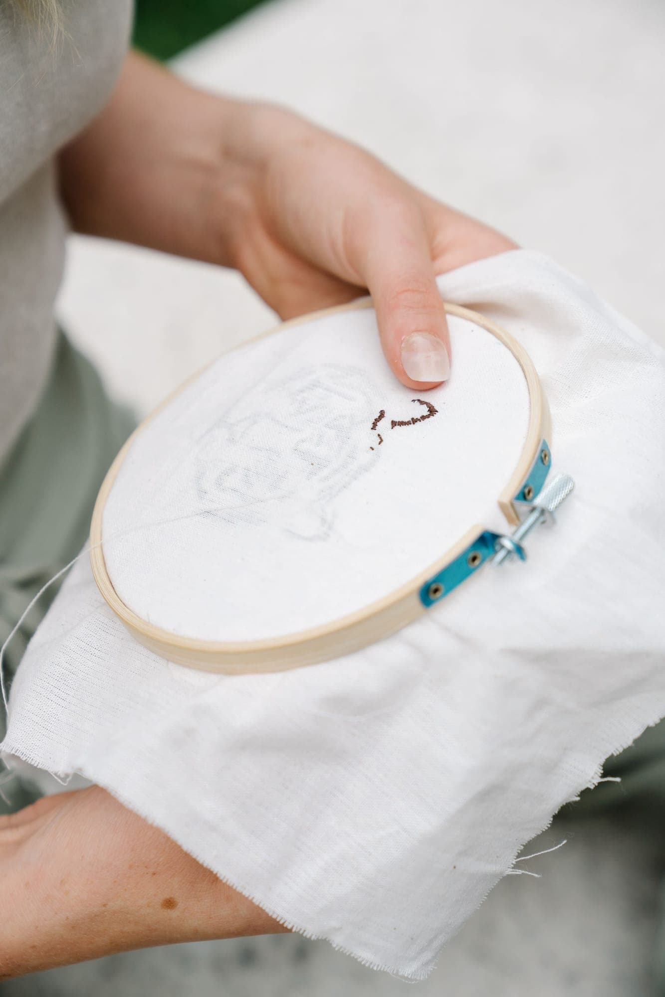Hands holding a wooden hoop with white fabric and the first stitches of a design, in daylight