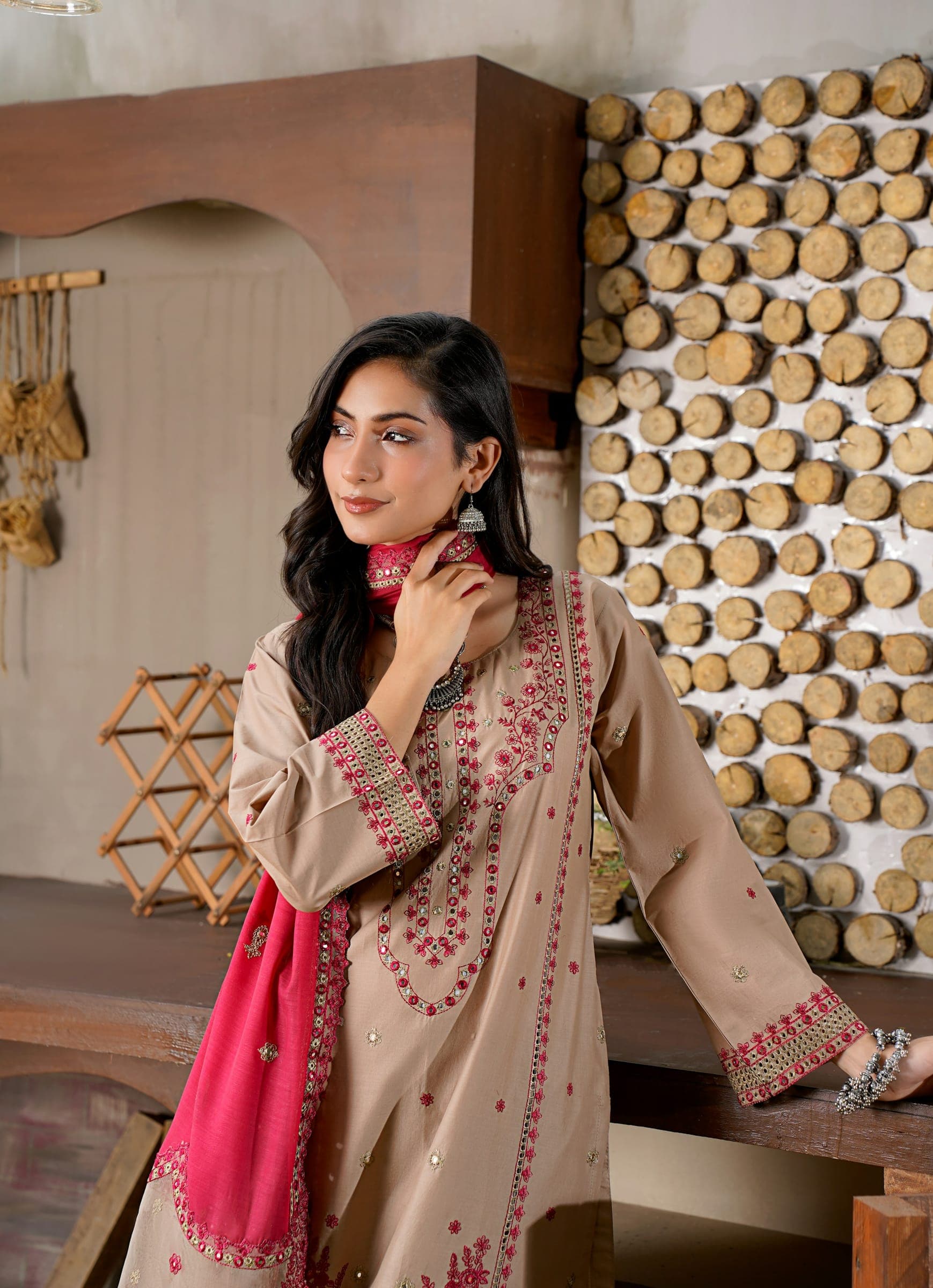 Woman in a lightly embroidered taupe kameez with a pink dupatta, standing by a wooden wall in daylight