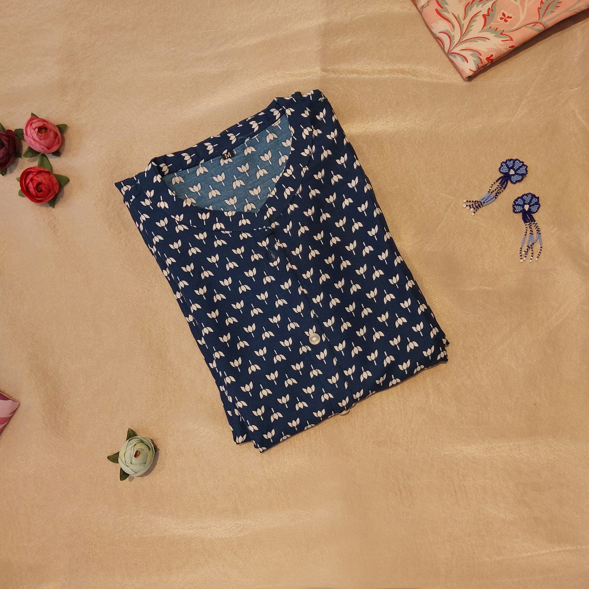A folded navy block-print cotton kurta on an ivory sheet with earrings and fabric flowers