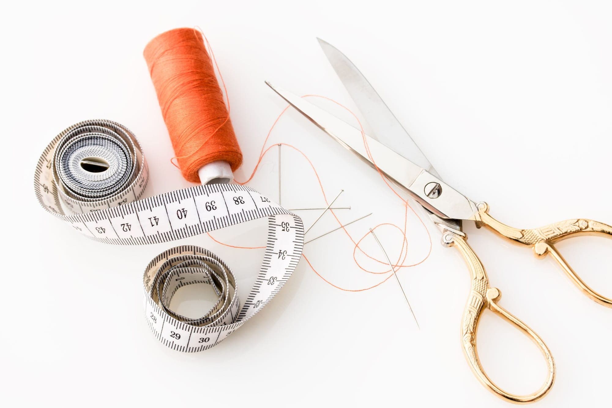 Gold-handled scissors, a tape measure, orange thread and a needle on a white surface