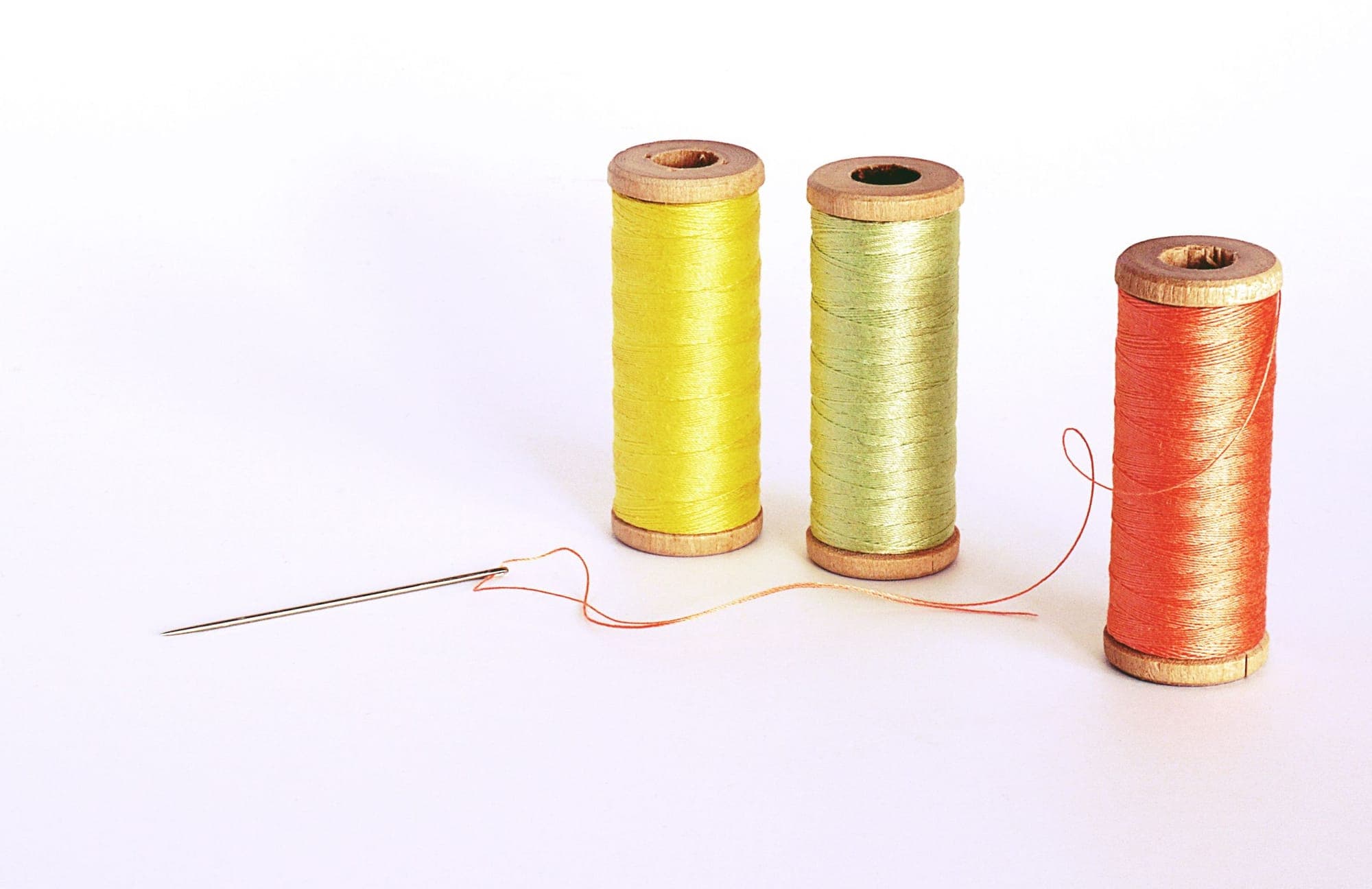 Three wooden spools of yellow, pale green and orange thread with a threaded needle on white