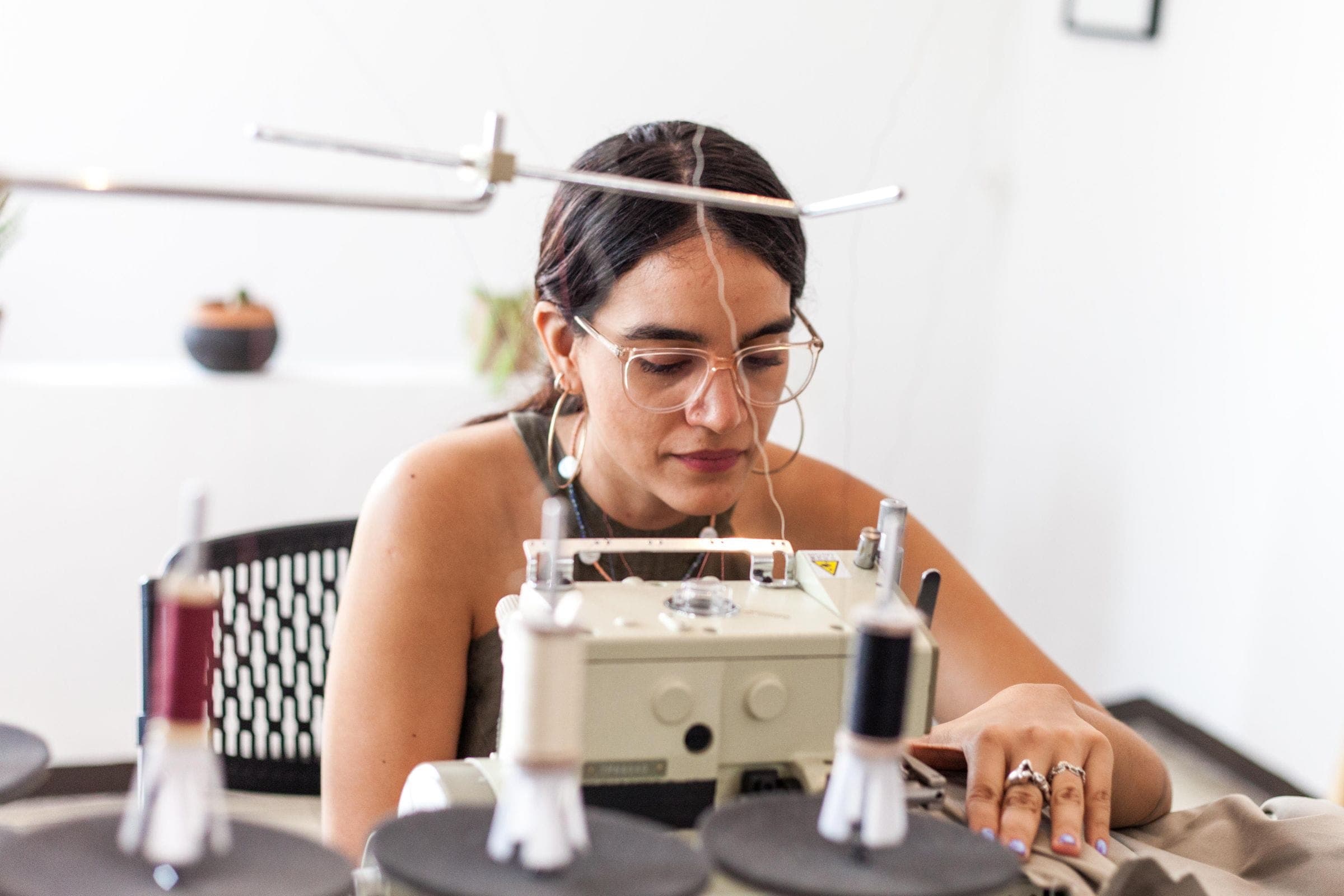 A woman stitching at a sewing machine in a bright studio with spools of thread in front