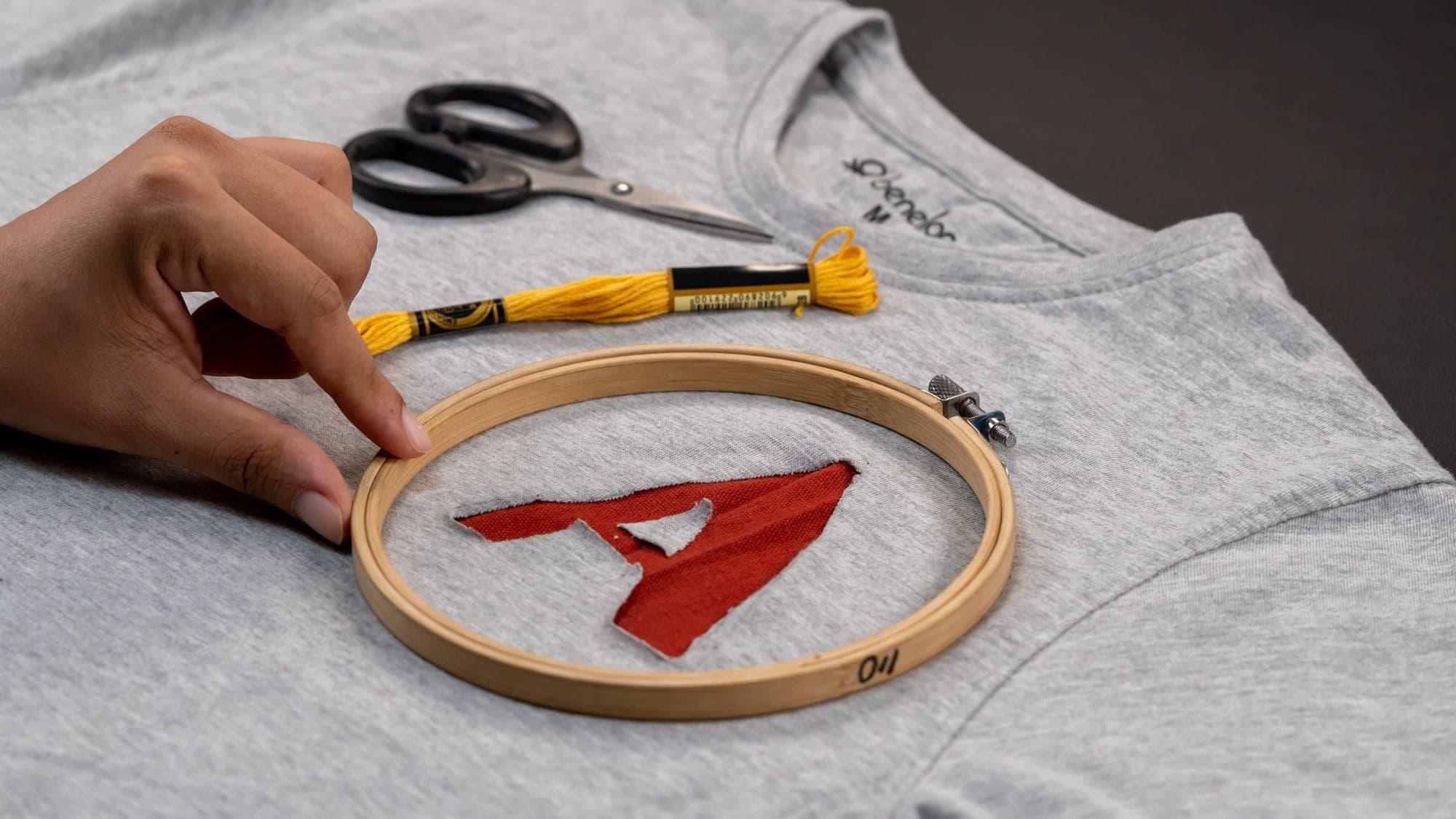 A red letter being embroidered in a hoop on a grey sweatshirt, with scissors and yellow floss